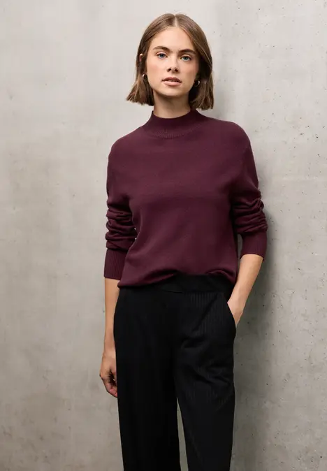 Frau mit bordeauxrotem Pullover und schwarzer Hose steht vor grauer Wand.