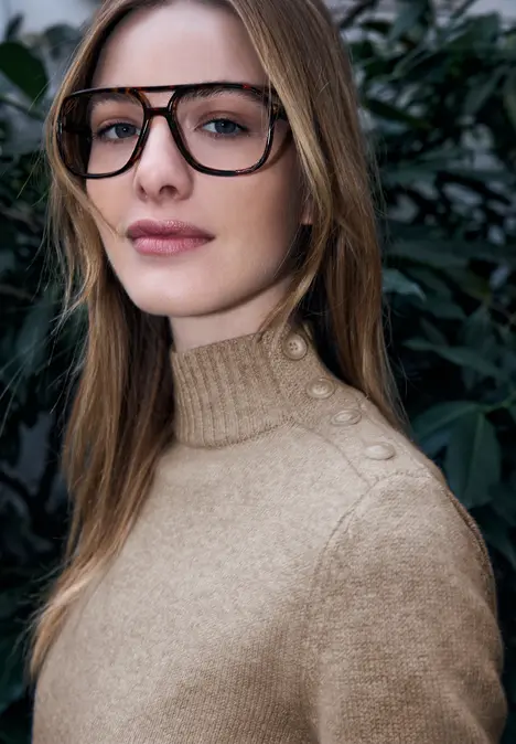 Frau im beigen Strickpullover mit Knöpfen an der Schulter vor grünem Laub.