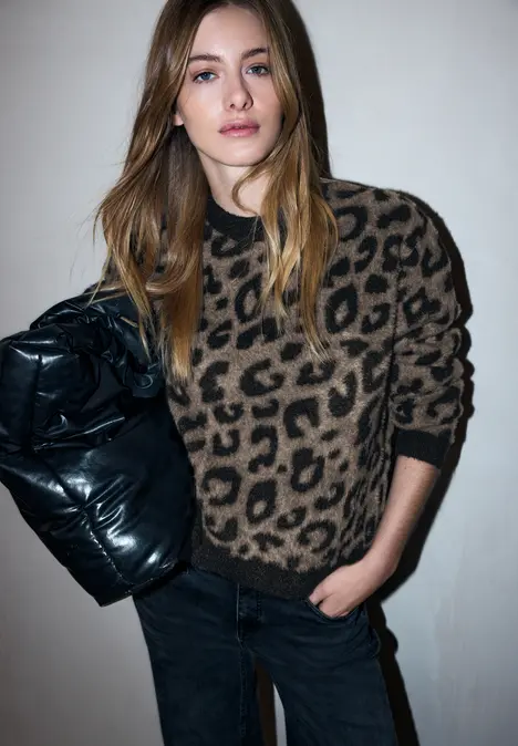 Person in Leopardenpullover und schwarzer Jeans hält eine schwarze Steppjacke unter dem Arm.