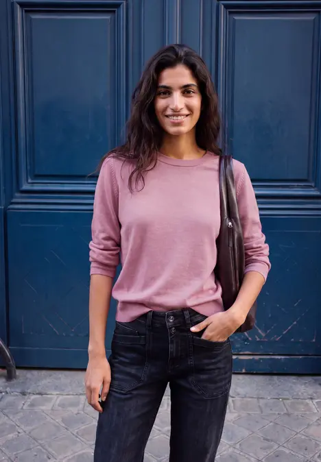 Eine junge Frau mit rosa Pullover und Jeans steht lächelnd vor einer blauen Tür.