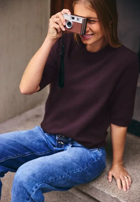 Frau sitzt auf einer Treppenstufe, trägt lila Shirt und Jeans und macht mit einer Kompaktkamera ein Foto.