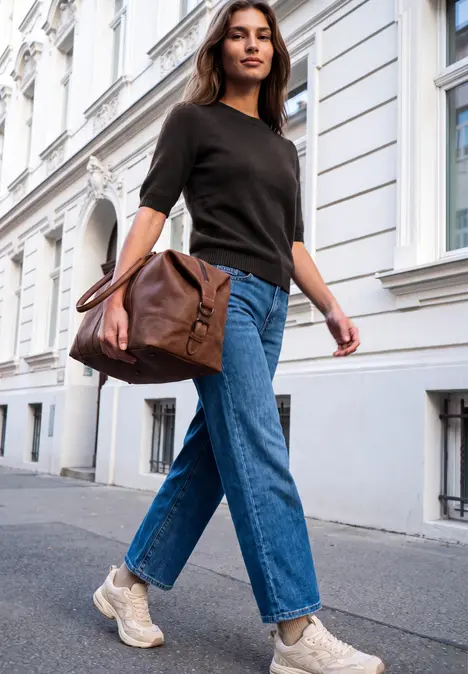 Junge Frau in Jeans, schwarzem Shirt und Sneakers trägt eine braune Ledertasche und geht die Straße entlang.