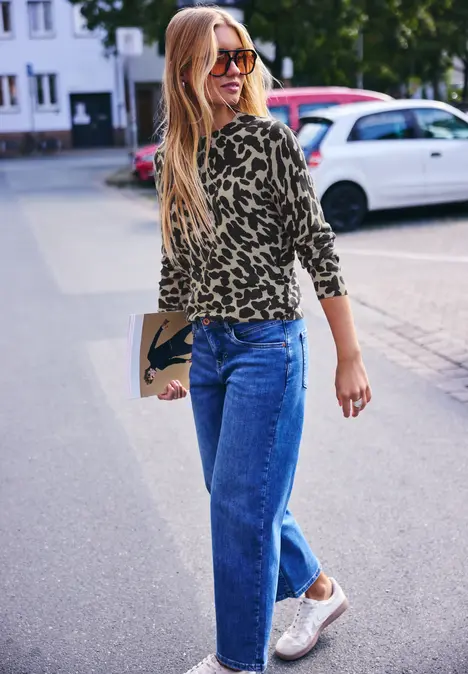 Eine Frau mit blondem Haar in Animal-Print-Shirt, Jeans und Sneakers läuft auf der Straße und hält eine Clutch.