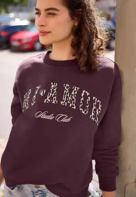 Junge Frau lehnt an einer Säule, trägt ein lila Sweatshirt mit floralen Buchstaben und Text 'Nature Club'.