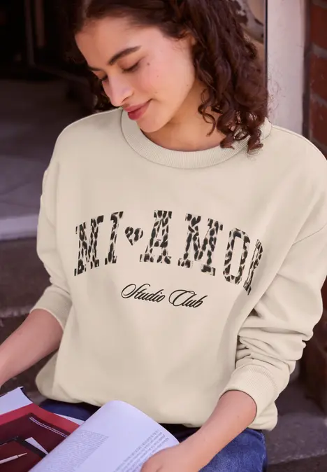 Junge Frau liest in einem Buch, trägt hellen Pullover mit Schriftzug Modern Club.