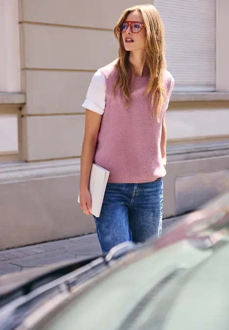 Junge Frau in rosa Strickpullunder, weißem T-Shirt und Jeans spaziert mit einem Laptop unter dem Arm.