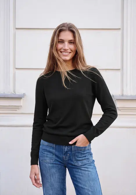 Junge Frau in schwarzem Pullover und Jeans, lächelnd vor heller Wand.