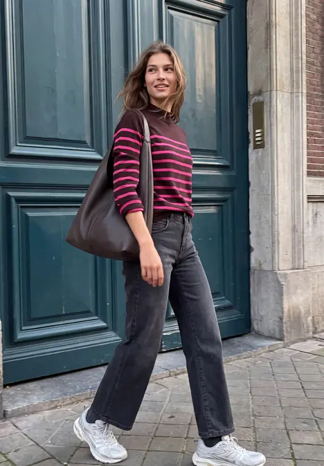 Une personne porte un pull rayé, un pantalon sombre et un sac à l'épaule, debout devant une porte.
