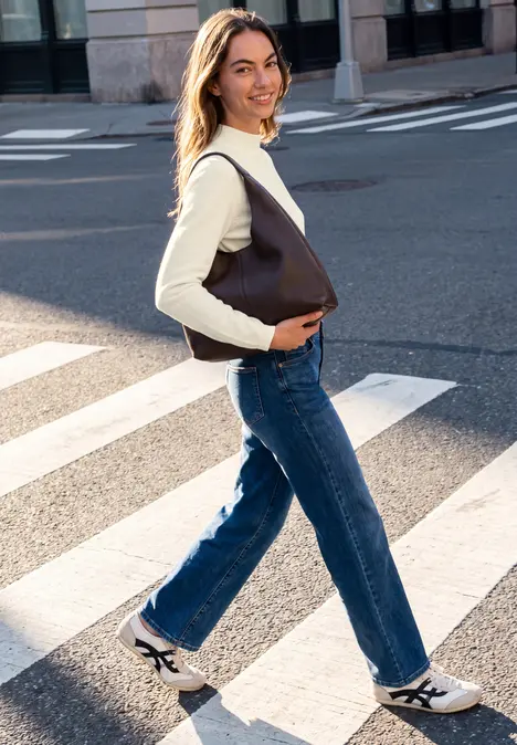 Eine junge Frau kreuzt lächelnd einen Zebrastreifen und trägt Jeans, Top und eine große braune Tasche.