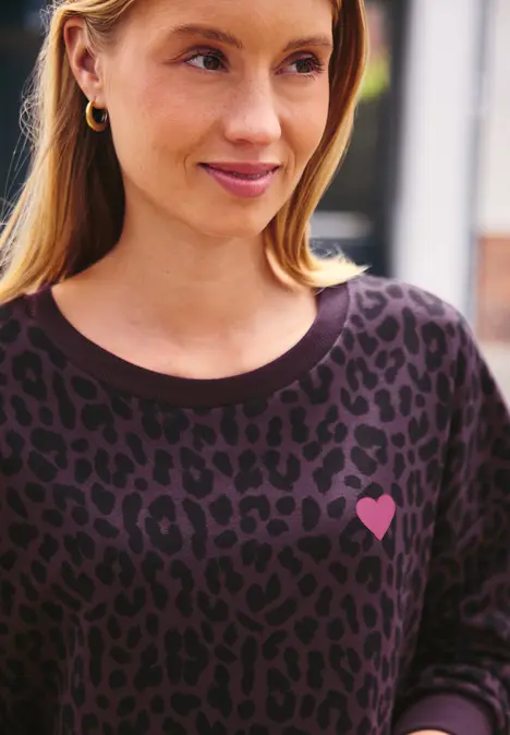 Frau mit blondem Haar trägt ein dunkelgeflecktes Shirt mit kleinem rosa Herz und lächelt leicht.