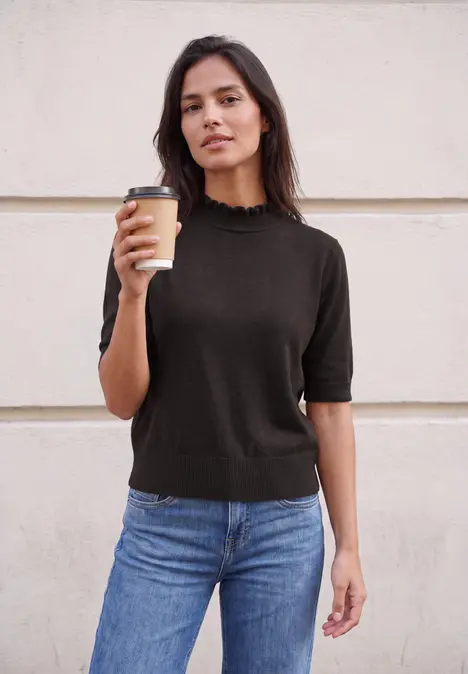 Frau steht vor einer Wand in schwarzem Pullover und Jeans und hält einen Coffee-to-go-Becher.