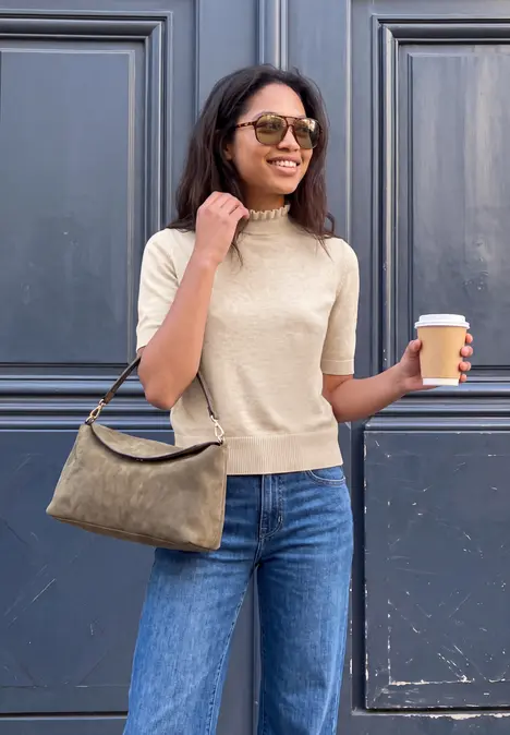 Frau mit Sonnenbrille, hellem Strickoberteil und Jeans trägt Handtasche und hält Kaffee.