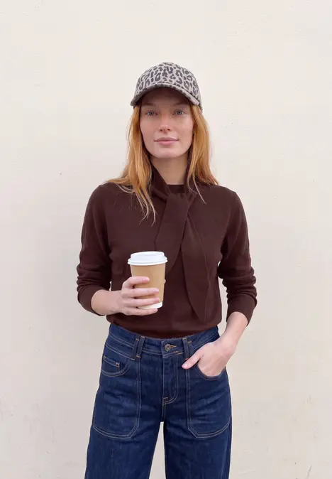 Eine junge Frau mit Cap und Kaffeebecher steht vor einer hellen Wand.