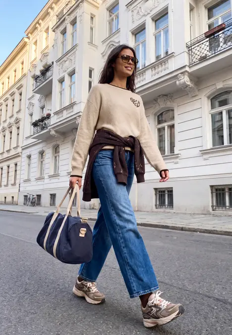 Eine modische Frau in Jeans, beigem Pullover und Sneakern mit Tasche spaziert über eine helle Stadtstraße.