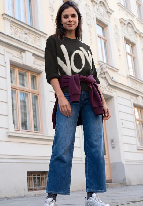 Eine junge Frau in Jeans und Pullover mit Aufdruck 'NOW' steht lässig vor einem Gebäude.