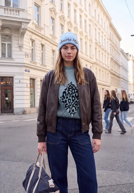 Junge Frau mit Jeans, Lederjacke und Leo-Pullover steht in Straßenszenerie.