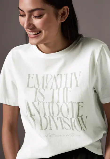 Frau mit weißem T-Shirt, Aufschrift: ‚Empathy is the antidote to division‘, sie lächelt.