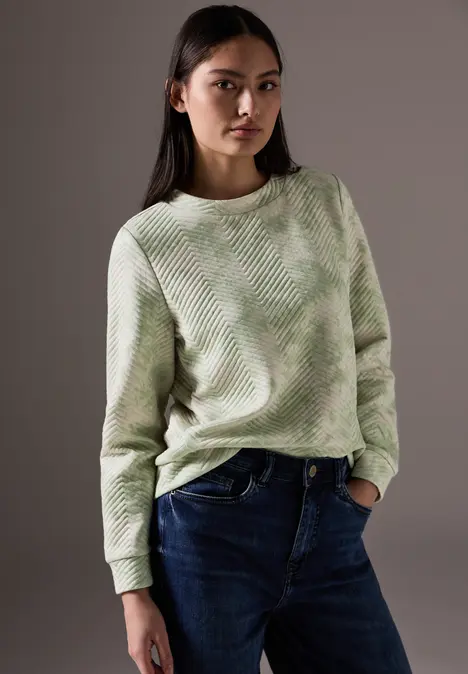Frau mit grünem Pullover und Jeans vor grauem Hintergrund.