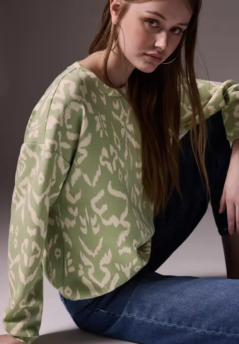 Frau mit grün gemustertem Pullover und Jeans sitzt am Boden.