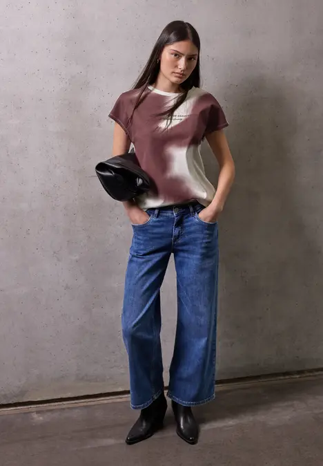 Frau sitzt entspannt auf Stuhl, trägt Jeans, Sneaker und gemustertes T-Shirt.