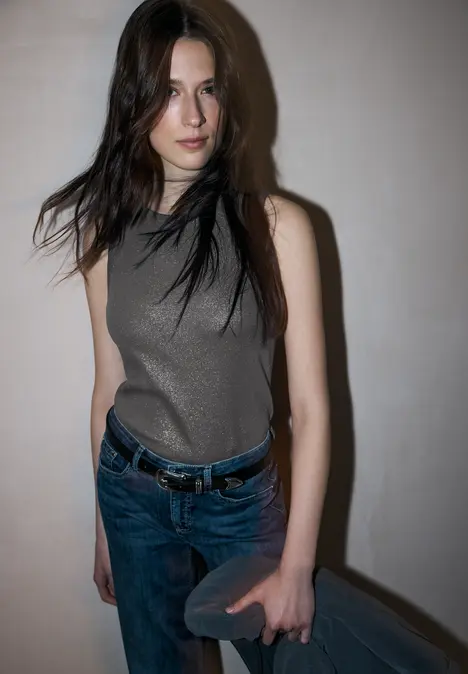 Frau in grauem Top, blauer Jeanshose mit Gürtel lehnt vor einer Wand und hält eine kleine Tasche.