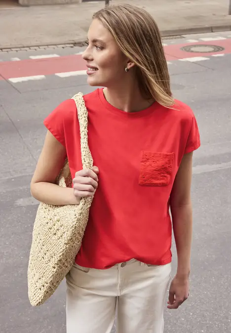 Eine Frau in rotem T-Shirt und weißen Hosen trägt eine beige gehäkelte Tasche und steht auf der Straße.