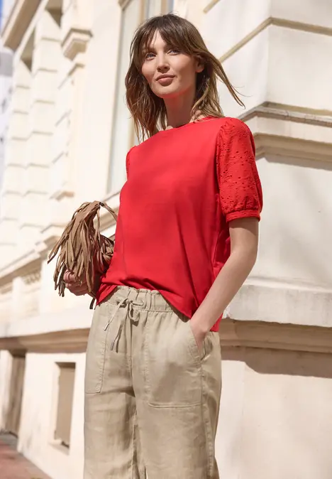 Frau in rotem T-Shirt und beiger Hose steht vor einem hellen Stadthaus mit Tasche in der Hand.