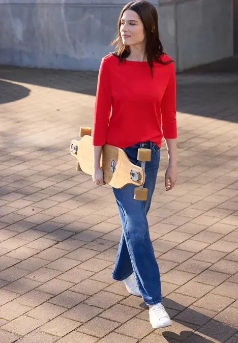 Frau in rotem Pullover und Jeans spaziert mit Skateboard entlang eines gepflasterten Wegs.