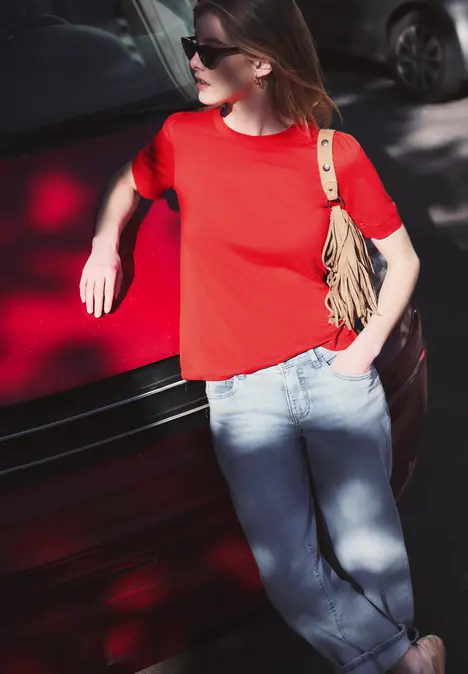 Frau im roten T-Shirt und blauen Jeans lehnt lässig an rotem Auto.