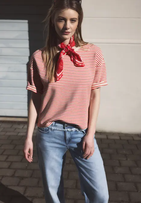 Eine Person steht vor einer hellen Hauswand, trägt ein rot-weiß gestreiftes Shirt, rotes Halstuch und blaue Jeans.