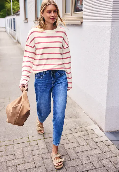 Frau in gestreiftem Pullover und Jeans trägt Papiertüte auf Pflasterweg
