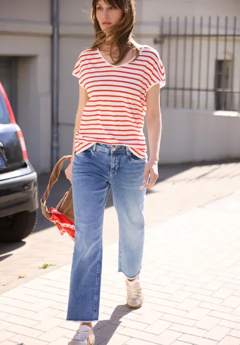 Frau in rot-weiß gestreiftem T-Shirt, weiten Jeans und Sneakern spaziert auf städtischem Gehweg.