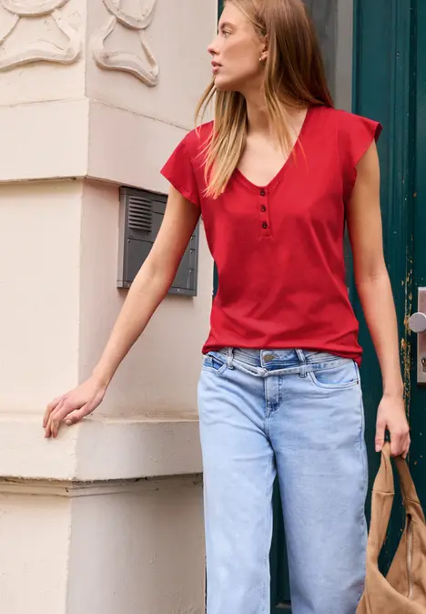 Eine Frau mit rotem Top und blauer Jeans, steht vor einem Gebäude und trägt eine Tasche (Gesicht unscharf).