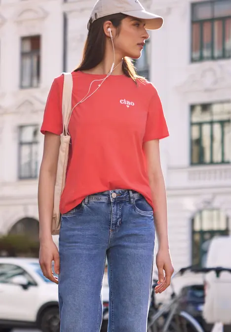 Junge Frau mit rotem T-Shirt, Jeans und Kopfhörern steht vor historischem Gebäude auf der Straße.