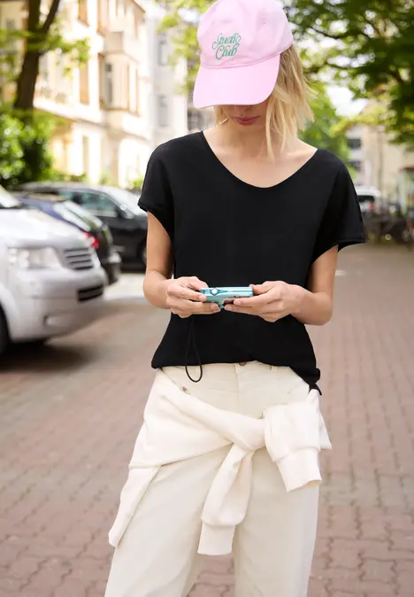 Person mit rosa Cap, schwarzem T-Shirt und hellem Pullover um die Hüfte, steht auf Straße und schaut auf ihr Handy.