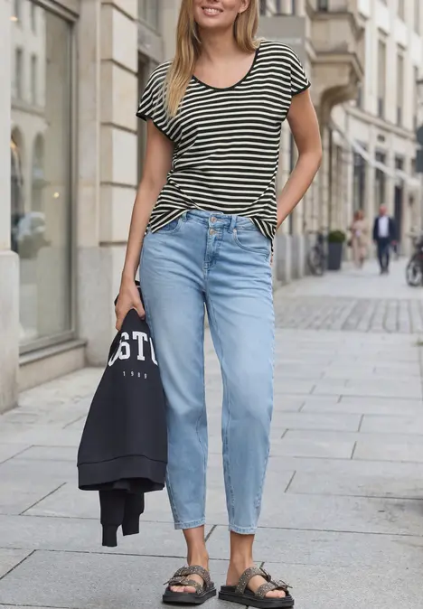 Eine Frau in gestreiftem T-Shirt, blauen Jeans und Sandalen spaziert in der Stadt und trägt eine Jacke in der Hand.