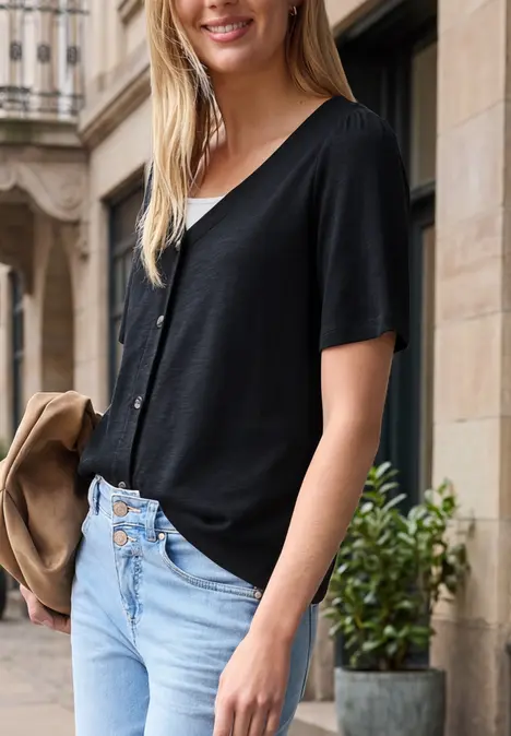 Frau trägt schwarzes V-Shirt und helle Jeans, lehnt an urbaner Fassade mit Pflanze im Hintergrund.