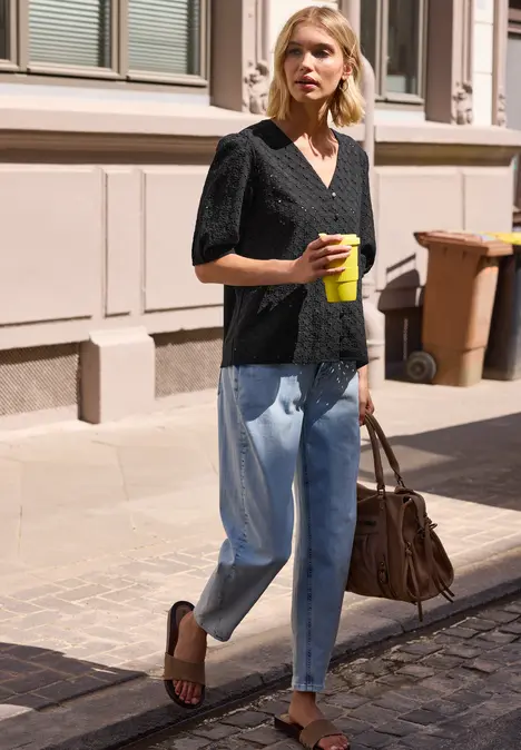 Eine Frau in schwarzer Bluse, Jeans und Sandalen geht mit Kaffeebecher und Ledertasche eine Straße entlang.
