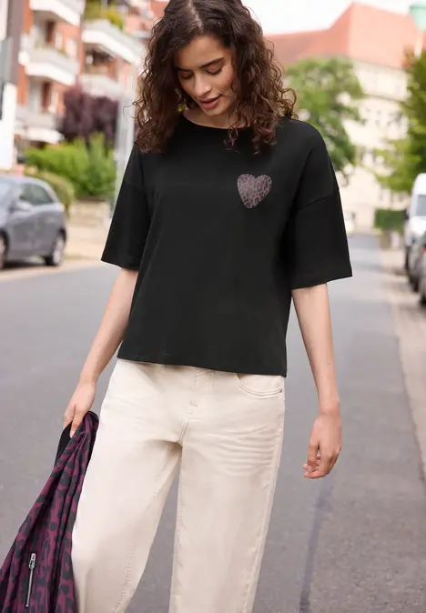 Frau in schwarzem T-Shirt mit Herz-Emblem und weißen Hosen spaziert auf der Straße, hält eine gemusterte Tasche.