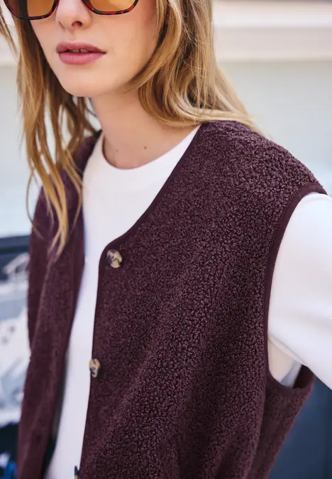 Auf dem Foto sieht man eine Frau in einem weißen Pulli und einer flauschigen, bordeauxroten Weste mit Knöpfen.
