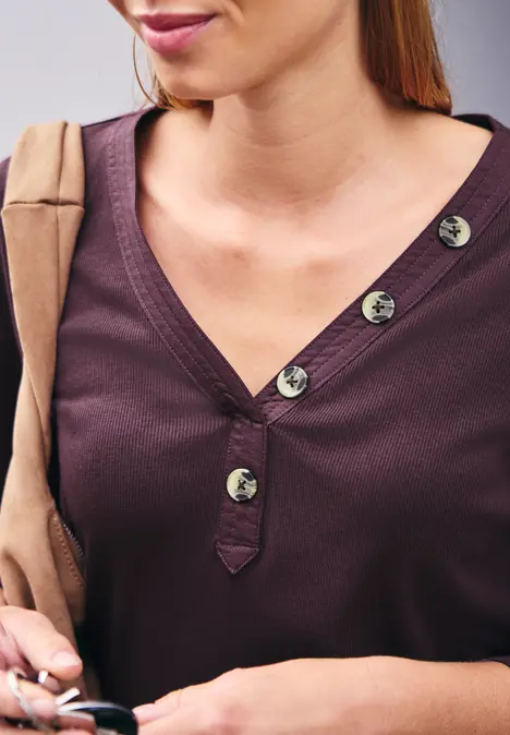 Frau in lila Oberteil mit silbernen Knöpfen trägt beige Jacke und hält Schlüsselbund.