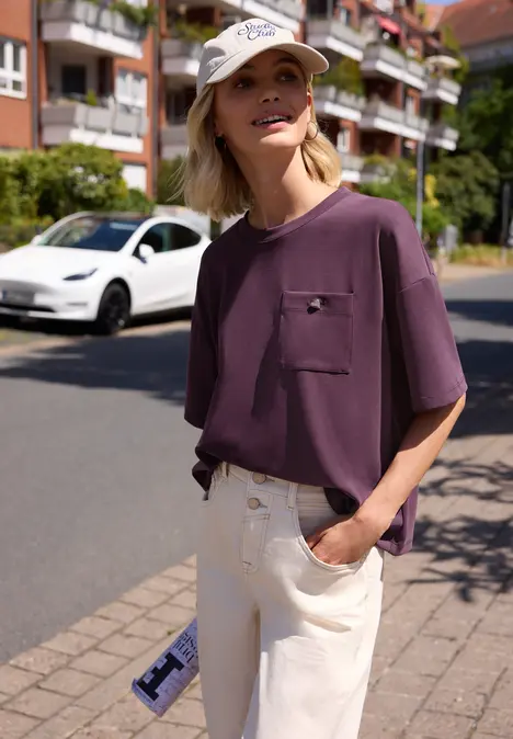 Person in lila T-Shirt und hellen Hosen steht entspannt mit Hand in Tasche auf dem Bürgersteig einer Wohnstraße.