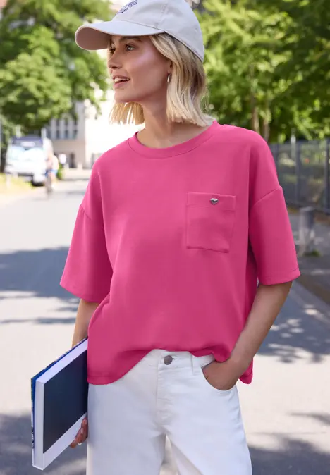Frau im pinken T-Shirt mit Tasche und weißer Cap, hält Tablet und steht im Freien.