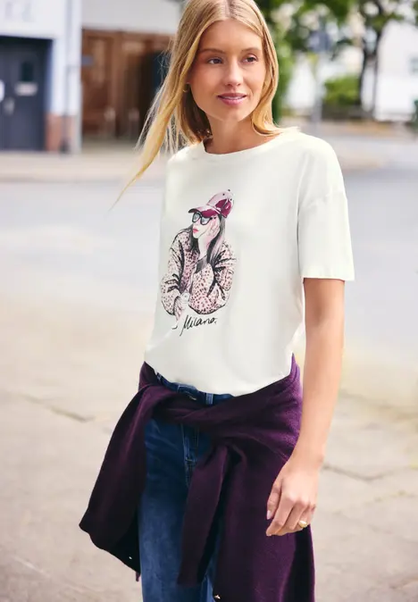 Junge Frau in weißem T-Shirt mit Katzenmotiv und lässigem Pullover um die Hüfte auf der Straße.