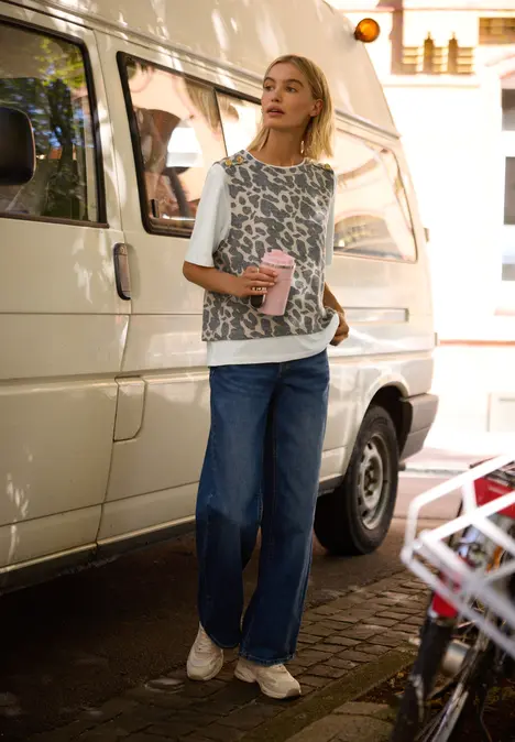 Eine junge Frau lehnt an einem weißen Van, hält einen Coffee-to-go-Becher und blickt nach oben.