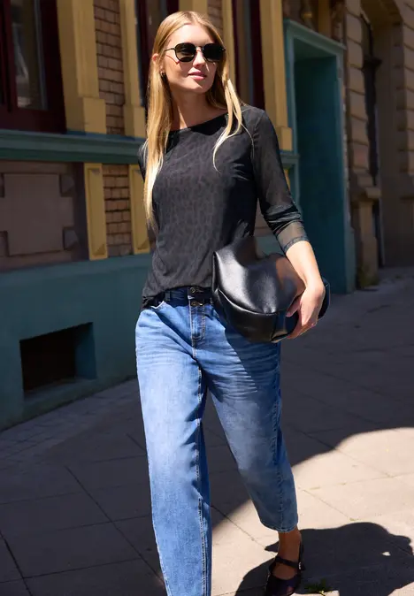 Junge Frau mit Sonnenbrille, schwarzem Top, blauen Jeans und Tasche spaziert sonnig die Straße entlang.