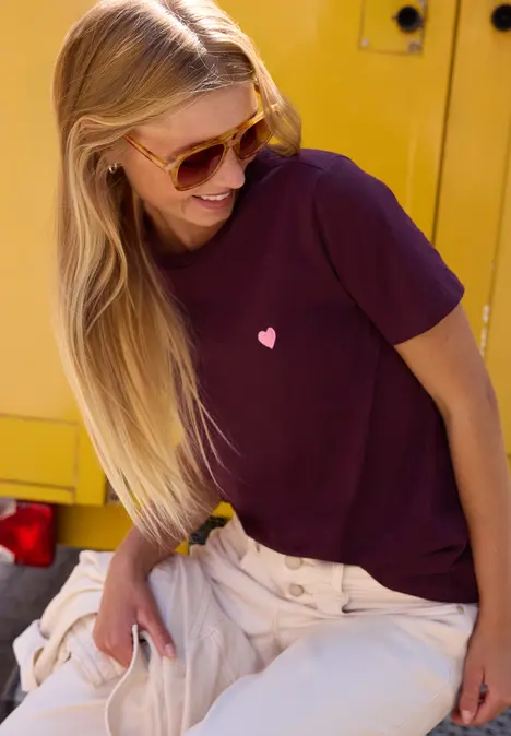 Lächelnde junge Frau mit Sonnenbrille und dunklem T-Shirt mit Herzmotiv vor einem gelben Fahrzeug.