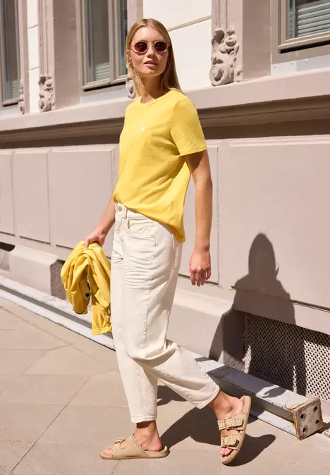 Frau in gelbem T-Shirt, heller Hose und Sandalen spaziert sonnig an einer Hauswand entlang.