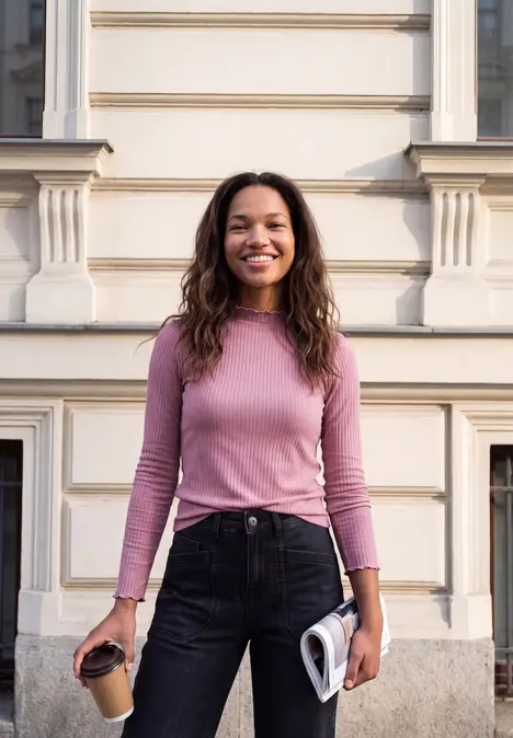 Jeune femme en pull rose et jean, souriante devant un immeuble, tenant un café et un magazine.