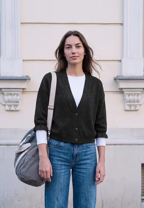 Junge Frau mit Cardigan, weißem Shirt, Jeans und Tasche steht vor einer hellen Hausfassade.